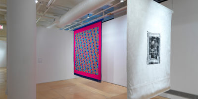 An Installation photograph of two large, ceiling to floor length, square textiles. The textile on the left has a thin, dark blue border with fringe draped from the bottom. Inside this border is another, much thicker, dark pink border. Inside is a zig-zag design of small squares in blue, red, teal, and orange colors. The textile on the right has a thick, white border. Inside the border is a smaller, black rectangular shape with white drawings of lines,dots, dashes and bolded lines.