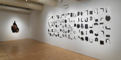 An installation photograph of the side walls of the gallery. On the right wall, four long rows of black shapes are painted on square papers. The four rows of paper extend horizontally across the entire wall. On the left wall is a three-dimensional sculpture made of brown yarn and other dark, found materials. The three-dimensional sculpture is in the shape of an upside-down heart