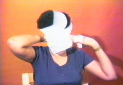 A film still of a middle aged person. The film still is of the person’s front profile from the shoulders up. Behind the person, is an orange wall. The person is in the motion of wrapping a white cloth around their face.