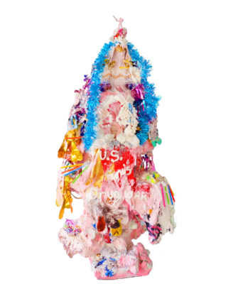 A photograph of a sculpture made of mixed materials in the space of a christmas tree. Shades of pink are painted on the base, and blue and gold colored garlands and lace are draped along its top and sides. On the top of the sculpture, a very small, pink figuring is standing.