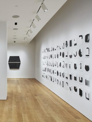 An installation photograph of the side walls of the gallery. On the right wall, four long rows of black shapes are painted on square papers. The four rows of paper extend horizontally across the entire wall. On the center wall to its left, there is a large, black sculpture of two trapezoids adhered to the wall.