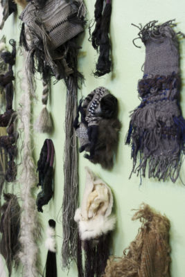 An installation photograph of the detail of a sculpture. Multiple found materials of textiles, hair and yarn are adhered to a green painted background. The green painted background is in the shape of a rhombus.