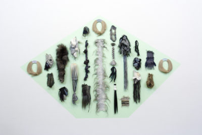 An installation photograph of multiple found materials of materials. Materials include textiles, hair and yarn. All materials adhered to a green painted background.