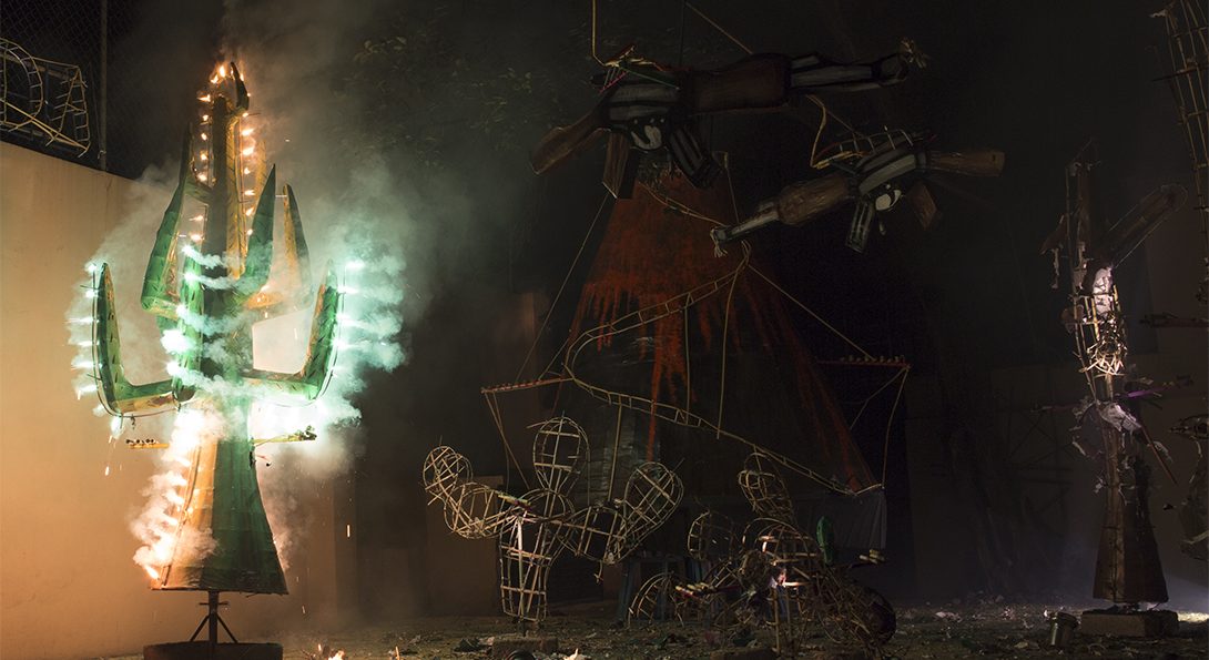 A photograph of a pyrotechnic performance at night-time. .A green tree sculpture is burning with a green glow. Behind the green- glowing sculpture is multiple, unlit, metal sculpture outlines.