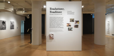 An Installation photograph of the gallery. A separate, square column stands on its own in the center of the photograph. On the column is adhered text of the exhibition information. The title, in large, bold texts, writes, “Traduttore, Traditore”. On the left wall, there are two large black and white, portrait photographs of middle-aged people.