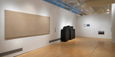 An installation photograph of two gallery walls meeting in a corner. On the left wall, there is a large, tan canvas. The canvas is about nine feet wide and 3 feet tall. To the right of the vancas is a large, four foot tall sculpture made of wood, painted entirely black. The sculture’s base is four large rectangles. On the right wall there are two images, eight by ten inches small. The pair of images are about two inches apart from each other. The image on the left is a mostly-white space in a black frame. The image, closely on the right, is of a landscape with a blue sky.