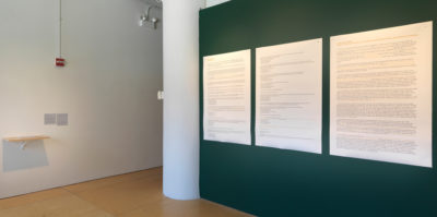 An installation photograph of two gallery walls. The wall on the left is white and the wall on the right is a dark green color. The wall on the left has a wooden shelf installed, with a small translucent paper placed on top. The dark, green wall on the right has three, large, archival pigment prints. The prints are arranged in the center of the wall in a horizontal line. Each print is about three feet tall and two feet wide. The color of the prints are white with small black text.