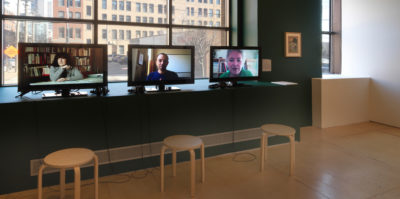 An installation photograph of three, small, computer monitors. The computer monitors are situated on a dark green painted wall’s window edge. The monitor on the left is a still of a middle-aged person, seated at a desk, talking. The center monitor is a still of a middle-aged person from the chest up, looking at the camera. The monitor on the right is of an older-aged person from the shoulders up, looking at the camera.