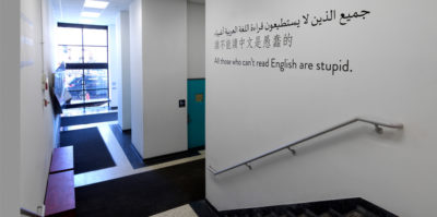 An installation photograph of the entrance outside of the gallery. The left side of the photograph is of stairs leading down to the ground floor. Ahead of the stairs is a hallway with black rugs leading to a ceiling to floor length window. On the right wall of the stairs, the sentence “All those who can’t read English are stupid.” is printed horizontally on the wall. The sentence is written in three different languages.