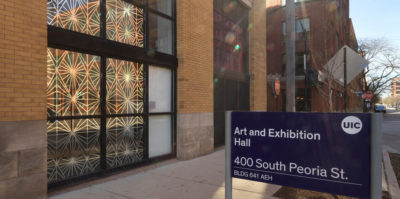 A photograph of the outside of a building. The building has two sets of three large windows between light-brown brick walls. Printed and displayed from the entire building window is a gold design of kaleidoscopic triangles. On the right side of the photograph, a blue sign reads “Art and Exhibition Hall, 400 South Peoria Street”