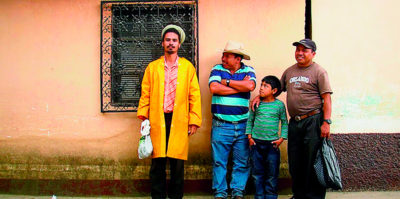 A photograph of three middle-aged people and one younger-aged person standing against an outside of an orange building.The person on the far left is wearing a long, yellow coat and a green hat. They are carrying a plastic bag. The three other people, to the person in the yellow coat’s left, are looking at them, smiling.