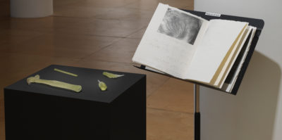 An installation photograph in the gallery space. On the left, a three foot tall, square podium is painted black. Placed on the podium are four sculptures made of sugar. The sculptures include replications of a hammer, a screwdriver, a pencil, and measuring tool. To the right of the podium piece is a black music sheet stand. An open book with a black and white photograph is placed on the stand.