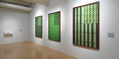 An installation photograph of two gallery walls. On the wall to the left, there is a shelf installation. On the shelf is an open book with red binding and white pages. The wall on the right side has three large, mounted, wooden-framed, ink-jet prints. The ink-jet prints are about the size of a standard american flag. Each print is a replication of the american flag, printed in variations of green shades.