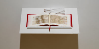 A photograph of an open book on a white shelf. The book has red binding and white pages. Printed on the pages are musical note sheets in black ink. A white glove sits on the end of the shelf.