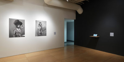 An installation photograph of two walls in the gallery. The left wall is painted white. Mounted on the left wall are two black and white, portrait photographs of middle-aged people. The size of the portraits are about three by two feet large. The right wall is painted black. Installed on the black wall is a white, thin shelf. Situated on the shelf is an iPad.