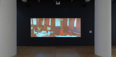 An installation photograph of a film room. Situated in the film room is a large projected film, paused on a film still. The film still captures two side-by-side scenes. The scene on the left is a group of five people sitting in a circle on the floor with a drum instrument. The scene on the right is one person sitting on the floor. Both scenes are in the same location of a room with red curtains.