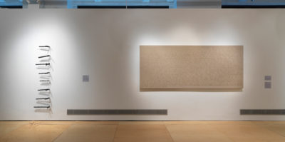 An installation photograph of a white gallery wall. On the left side of the wall, eight three-dimensional, thin objects are adhered in a vertical line. On the right side of the wall, there is a large, tan canvas. The canvas is about nine feet wide and 3 feet tall.