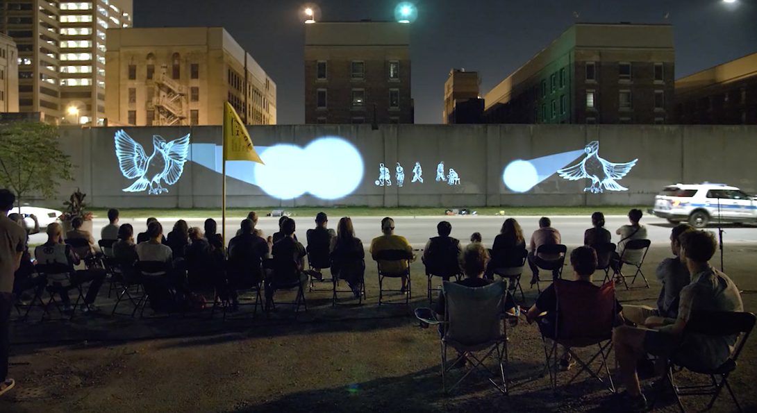 A photograph taken in the evening of about 28 people. They are sitting in lawn chairs, watching a projection against a concrete wall on the side of a highway.