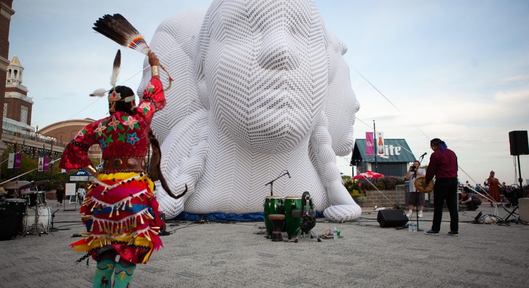 A large white sculpture of three heads, the size of houses, is placed in a courtyard. A young person is dancing in front of the sculpture. Next to the sculpture is a person playing an instrument.