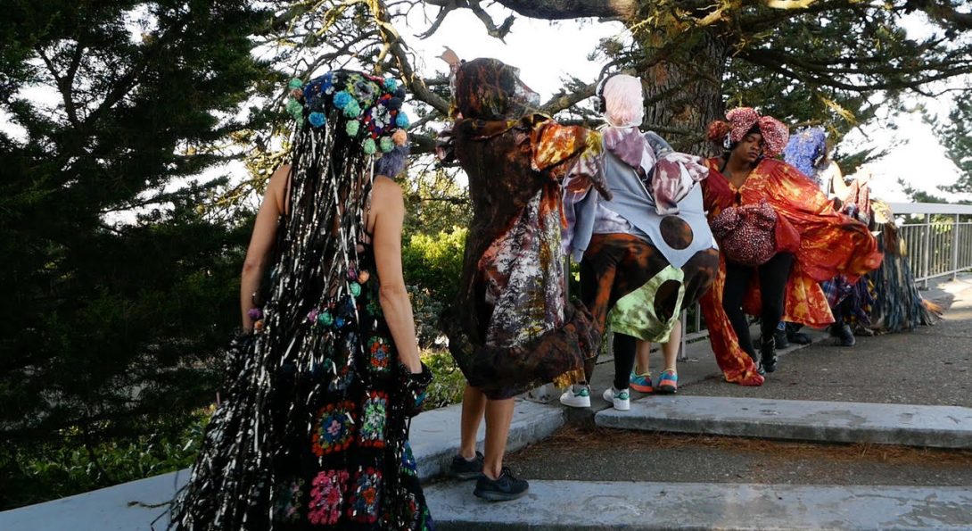 A photograph taken in landscape mode in a park captures three people standing in a vertical line next to one another. In the center, a person with orange patterned clothing with a bonnet-like hood looks down. They are all wearing silent disco headphones.