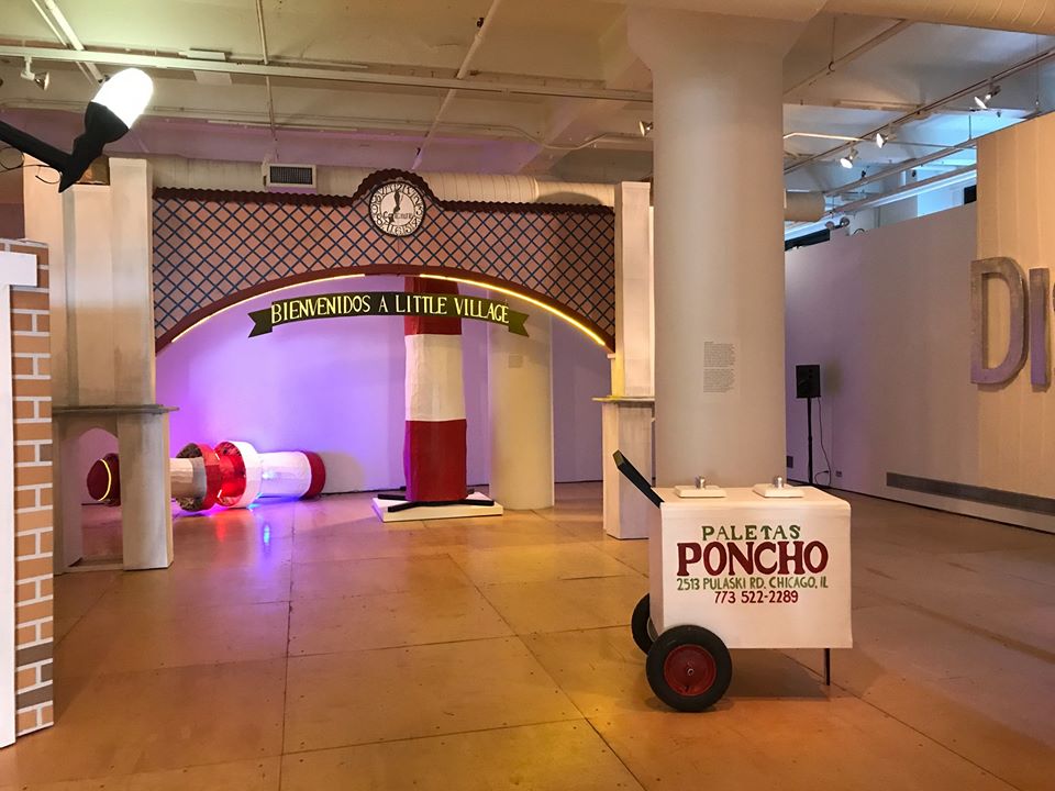 An installation photograph of The Last Judgment. This photograph is taken of the main gallery, in the center a large, maroon and brown arch sculpture that reaches from the floor to the ceiling. A white clock is centered in the top-middle point of the arch, below a ribbon design reads “Bienvenidos A Little Village”. In front of the arch, another sculpture, replicating a paletas cart sits on the right side of the photograph. The white paletas cart with red and green text and reads “Poncho, 2513 Pulaski RD, Chicago, IL”