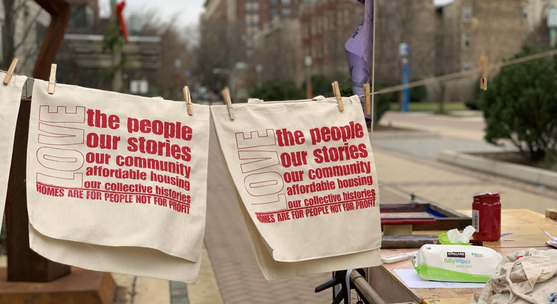 A photograph of two beige textiles with bold, red text printed over top. The textiles are displaced on a clothing pin string in an outside area. The red bold text writes, “LOVE the people, our stories, our community, affordable housing, our collective histories. Homes are for people, not for profit”