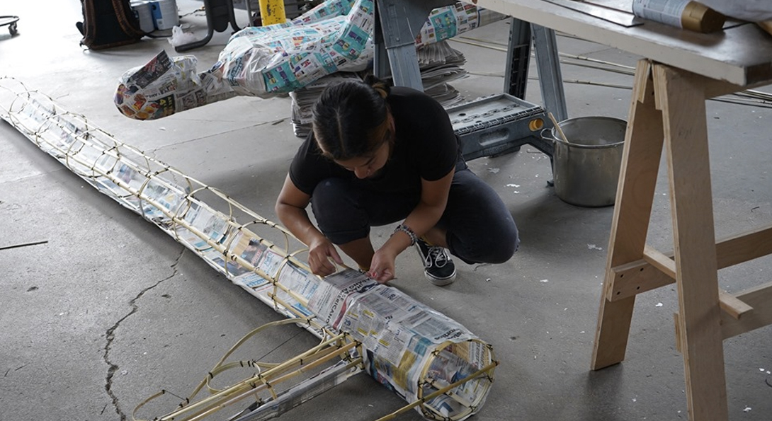 A photograph of a middle-aged person crouched onto the ground. They are working on long, circular sculpture with materials of wood and newspaper