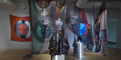 A dress form stands in the middle of a room filled with textiles hung from the ceiling. On the form is a denim outfit with a billowing skirt made of flannel scraps.