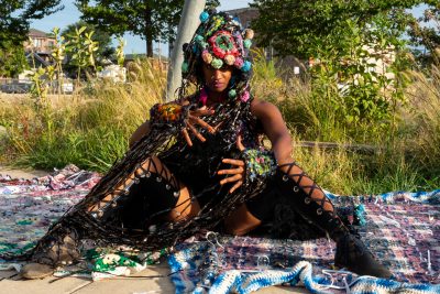 A dark skinned person in an elaborate costume sits in a natural area with their legs spread.