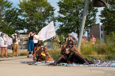 A dark-skinned person in black kneels and holds an object up towards the sky. Onlookers observe from the background.