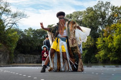 A dark-skinned person wearing a mask of fringe is caught mid-dance. A young girl looks on from the background.