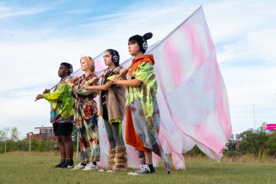 Four young people stand in a line, each are carrying a white and pink flag over their shoulder.