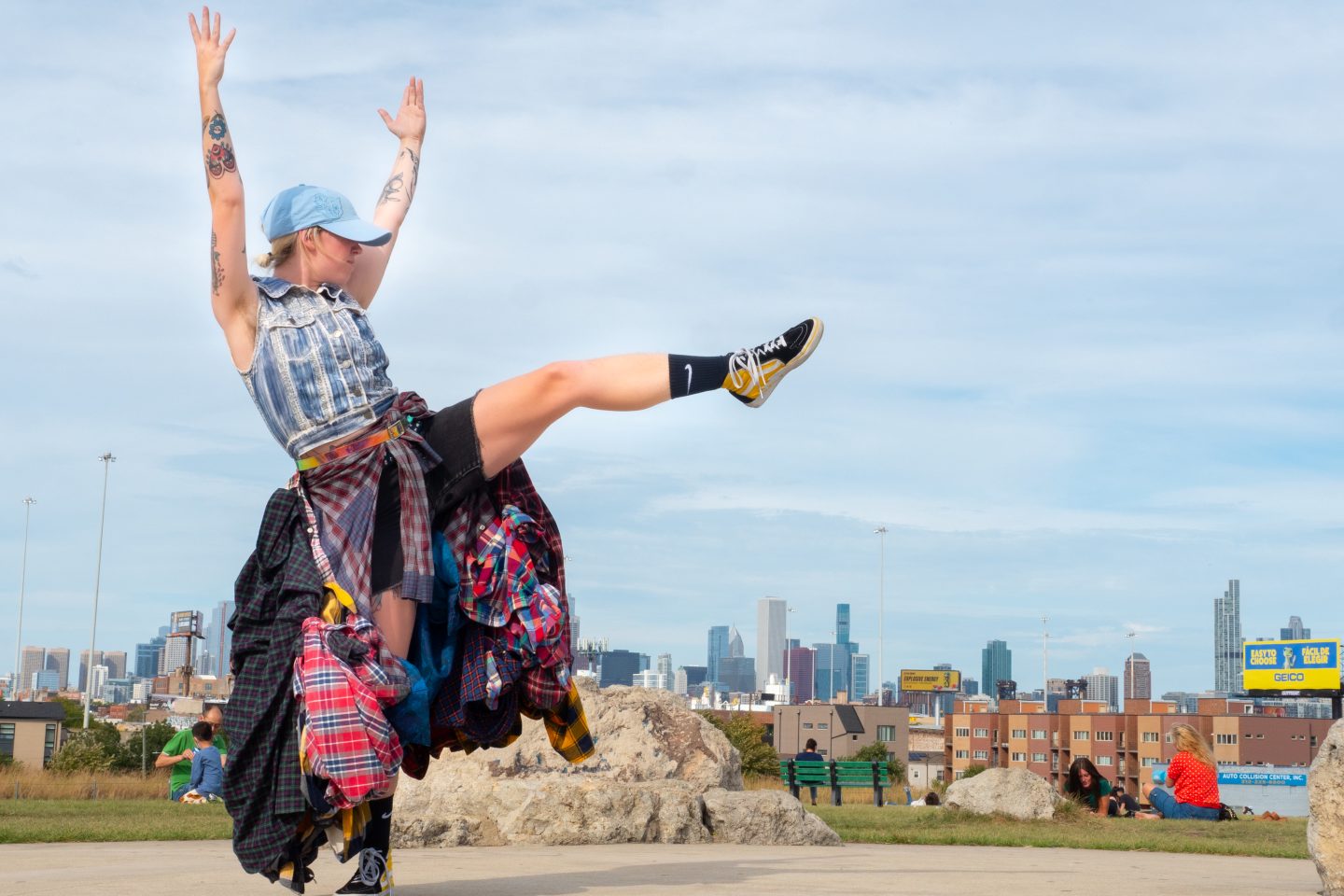 A light-skinned person in a baseball cap and skirt made of many layers of flannel, raises her leg, mid-dance.