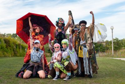 Seven people in colorful costumes stand and kneel in a group. They each have their fist raised.