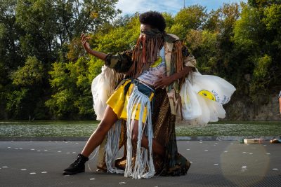 A dark-skinned person wearing a fringed costume and with many paper bags attached to the arms is caught mid-dance.