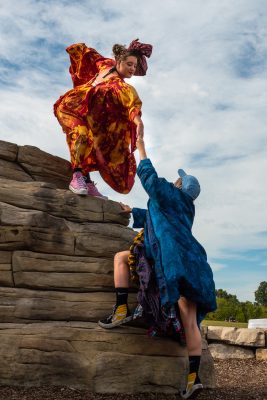 One person in an orange costume stands at the top of a rock. They reach down towards a person in blue, grasping their hand.