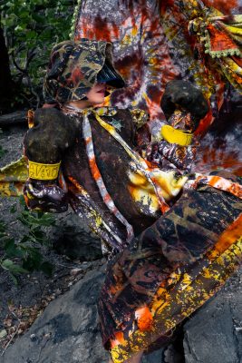 A light-skinned person in a brown and orange tie-dyed costume takes a boxer's stance. Only their mouth is visible under their hood.