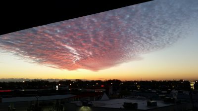 A long silutohtetre triangle silhouette with the right angle in the upper-left corner. Purple-pink cirrocumulus clouds above the town landscape, starting in the middle of the image then spreading out upwards in a roughly triangular shape, with red-pink highlights on the bottom and dark purple shadow on top from the sunset. The sky around the clouds fading from orange on the horizon at the lower quarter of the image to slightly medium dark blue upwards. Below the horizon are buildings in shadow with lights on and trees along the horizon line.