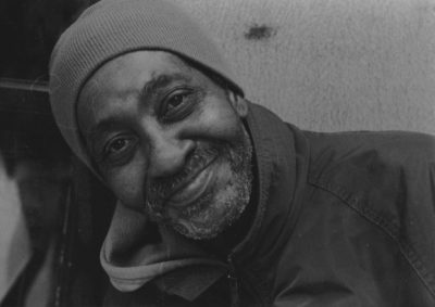 A black and white photograph, of a close-up shot of a beard, likely-black, man tilting his head to the left of the image in a beanie hat and a coat giving a closed-lipped warmth filled smile, facing towards the viewer.