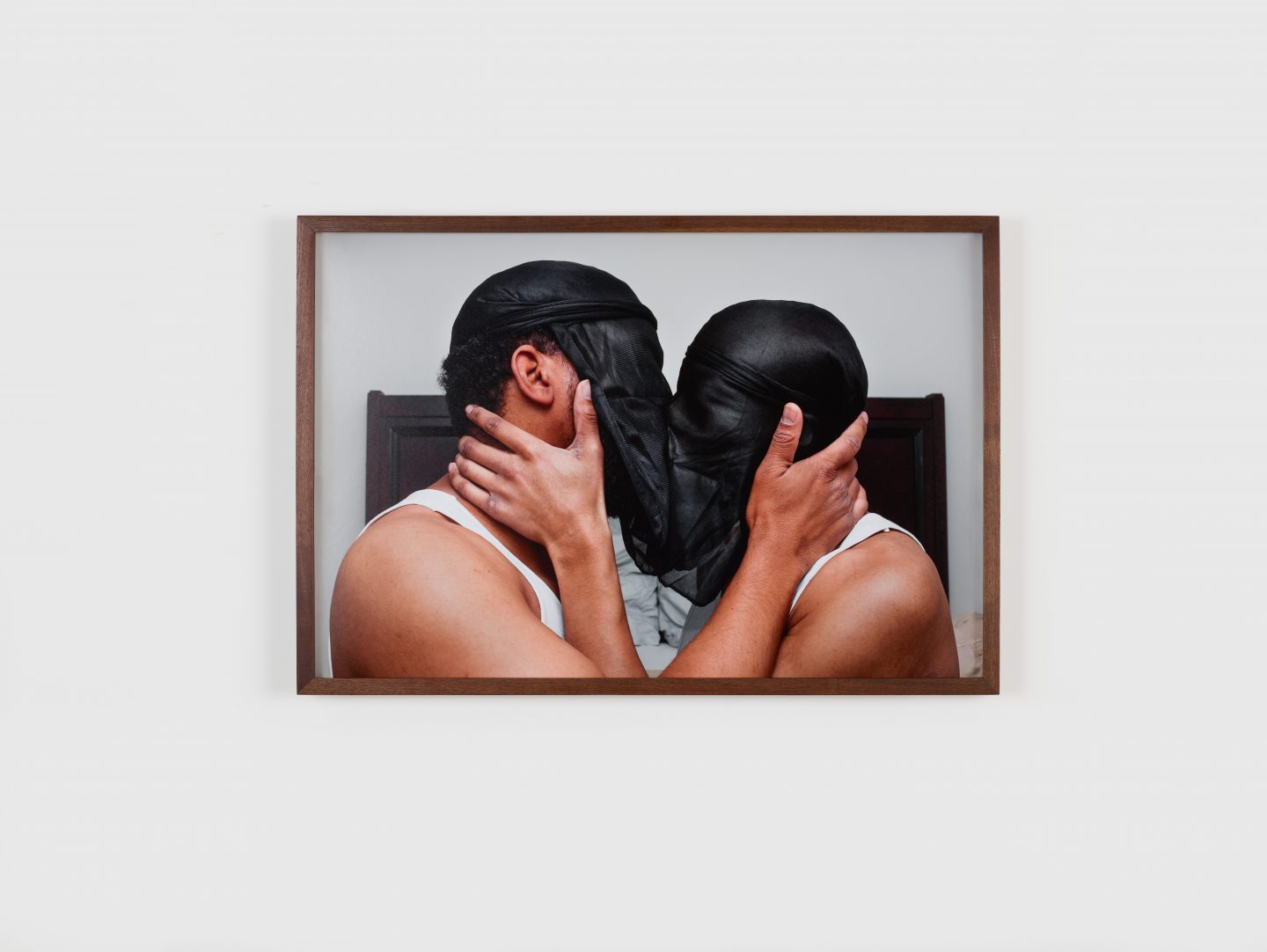 A photograph of two dark-skinned individuals kissing passionately in front of a white wall with their heads and faces covered by reversed black Du-Rags.