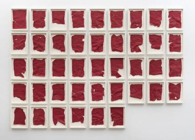 On a white wall, a number of lined up frames contain scraps of red fabric-like paper.