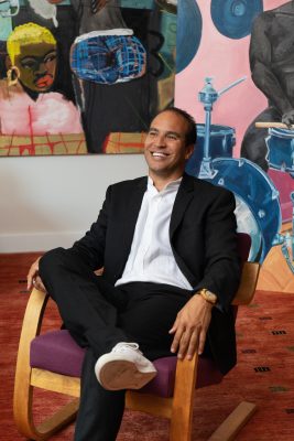 A black man in a suit sits in front of a contemporary art piece and smiles towards just off camera