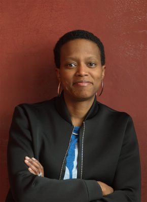 A black woman with short hair crosses her arms and looks into slightly to the left of the camera with a small smile on her face.