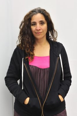 A woman with light brown skin and curly brown hair looks into the camera with a small smile.