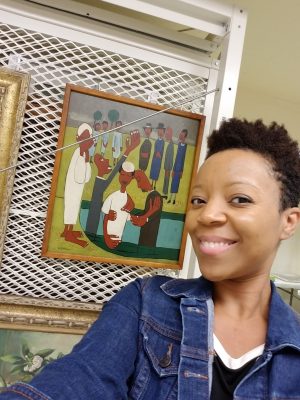 A black woman with short hair stands smiling in front of a painting.