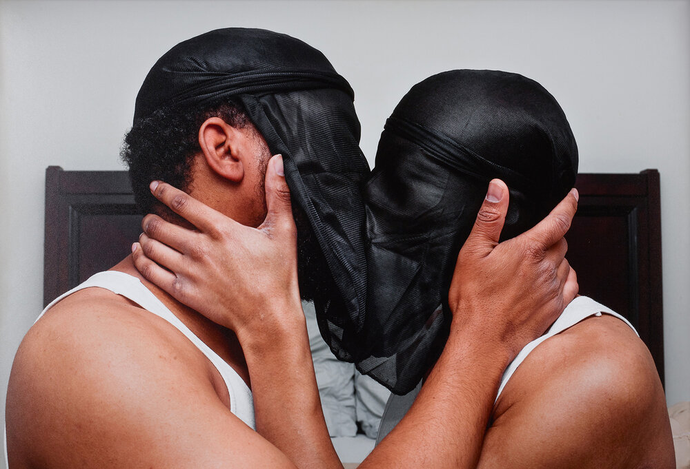 Two black men wearing black doo-rags around the front of their faces kiss while holding each other's faces.