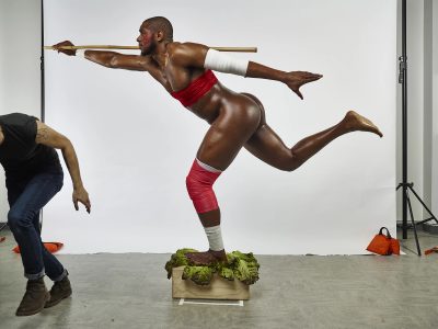 A black man wearing only strips of red and white fabric around his arms and legs stands on a wooden box in front of white screen. He is posed on one leg, facing towards the left and holding a wooden stick. There is a man in jeans and a teeshirt leaving the frame on the left.