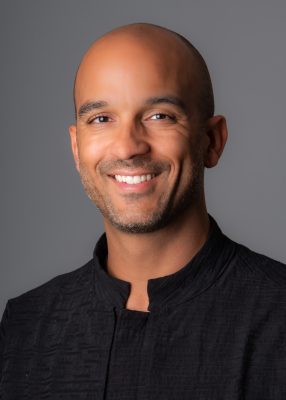 Headshot of a Black man smiling into the camera in front of a grey backdrop.