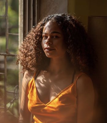 Headshot of a young Black woman wearing a yellow shirt and standing in front of a window.