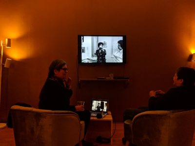 Two people sitting with their backs to the camera. They are in conversation and there is a tv with black and white footage on it. The rest of the photograph is orange tinted.